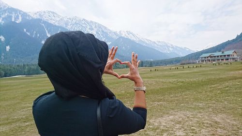 Rear view of woman making heart shape from hands on field against sky