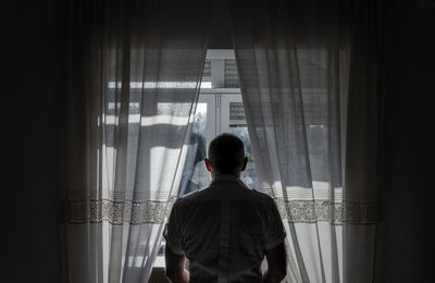 Rear view of adult man standing beside window with sunlight and shadow