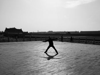 Man with arms outstretched standing on footpath against sky in sunny day