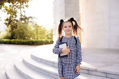 Sweet smiling child girl 5-6 year old wear checkered black and white dress and backpack hold book