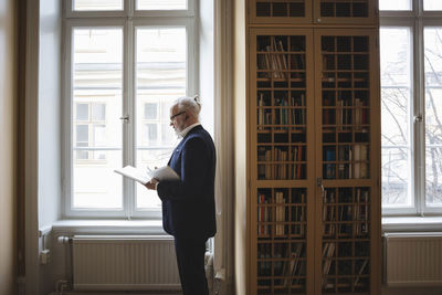 Senior professional reading book while listening through headphones in law library