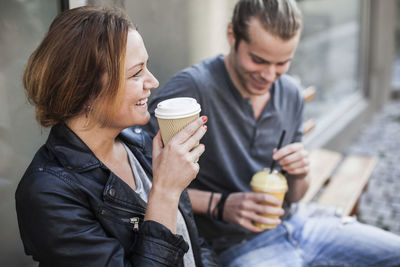 Happy woman drinking coffee in disposable cup with man on bench at sidewalk