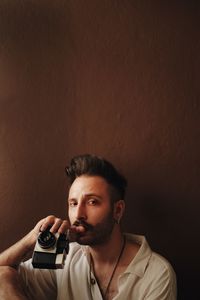 Portrait of young man holding camera against wall