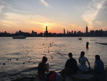 Friends sitting on rock formation at riverbank against skyline during sunset