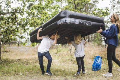 Full length of teenage girl having fun with siblings while carrying inflatable mattress together at camping site