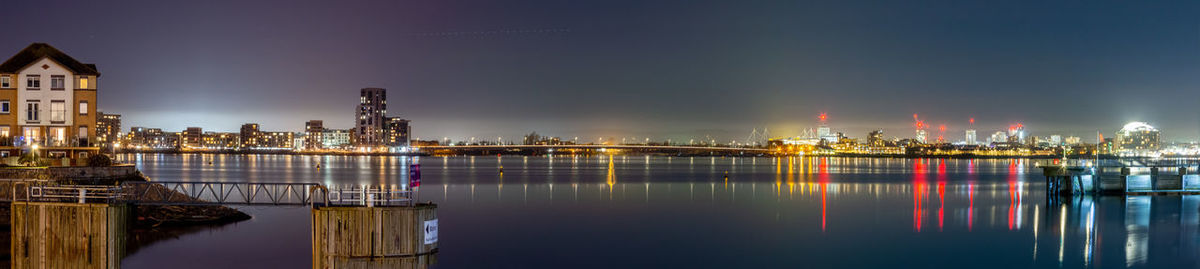 Illuminated buildings by river against sky at night