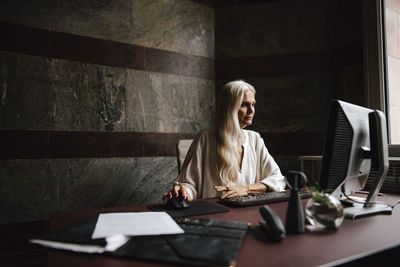 Confident mature businesswoman with long white hair using computer at desk in office