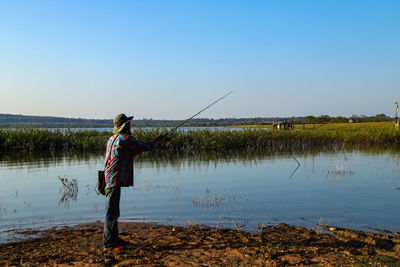 Rear view of person standing by lake against clear blue sky