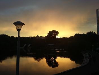 Illuminated street light by silhouette trees against sky at sunset