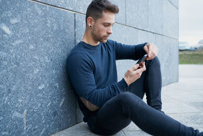 Side view of modern male in casual wear sitting near stone wall and br