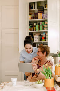 Smiling elderly woman sharing digital tablet with young female caregiver working in kitchen at nursing home