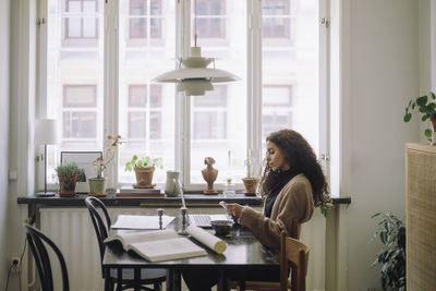 Side view of female architect using smart phone while sitting with laptop on table at home