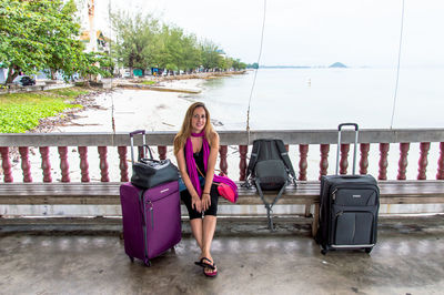 Full length portrait of beautiful woman with luggage sitting on bench against sea