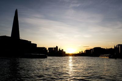 Silhouette buildings by river against sky during sunset