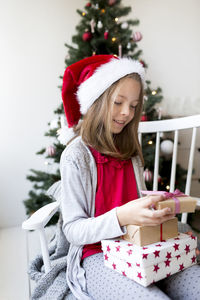 Smiling girl holding gift box while sitting at home against christmas tree