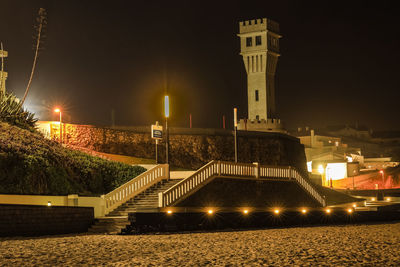 Illuminated street by buildings against sky at night