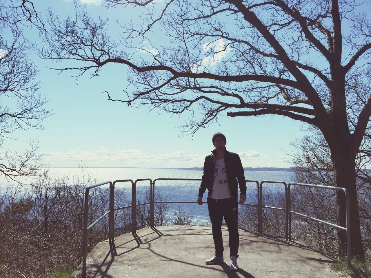 lifestyles, leisure activity, rear view, full length, water, railing, sky, standing, tree, tranquility, nature, casual clothing, tranquil scene, scenics, sea, branch, bare tree, beauty in nature