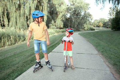 Caucasian boys brothers in helmets riding roller skates and scooter on road in park