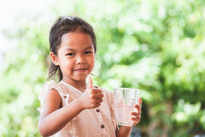 Portrait of girl gesturing thumbs up sign while holding water glass outdoors