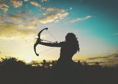Silhouette teenage girl aiming archery against sky during sunset