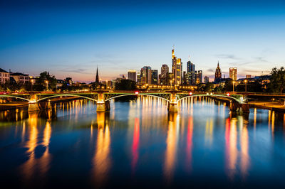 Illuminated bridge over river by buildings against sky at dusk