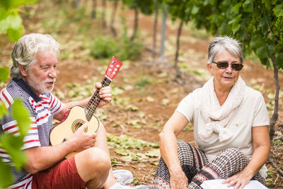 Senior man playing guitar while sitting by woman at vineyard