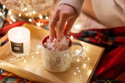 Woman's hand applies marshmallows on cup of cocoa on tray on red plaid and garland of glowing lights