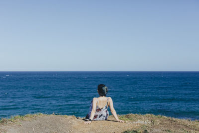 Rear view of woman sitting on cliff by beach against clear sky