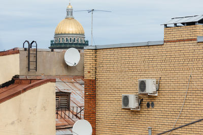 Two white air conditioners mounted on yellow brick wall and two sattelite dishes for television. 