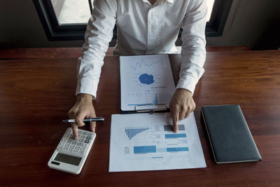 Midsection of man analyzing chart with calculator while sitting at office desk