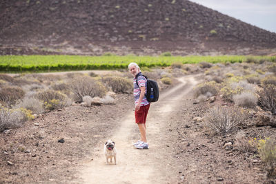 Side view of man hiking with pug on dirt road against mountain