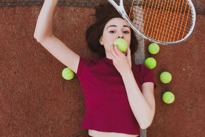 High angle portrait of woman holding tennis racket and ball while lying on court