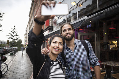 Couple taking selfie through smart phone while standing by sidewalk cafe in city