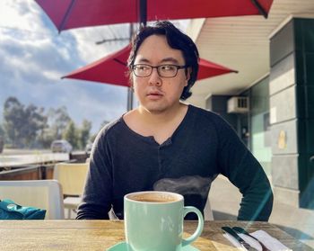 Young asian man having hot coffee at sidewalk cafe under red cafe umbrella.