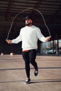 Full body of active bearded male in black cap jumping rope while training on sunny street under roof n city