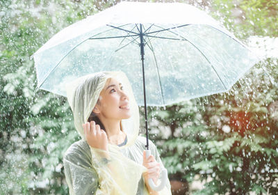 Young woman holding umbrella while standing against plants during rain