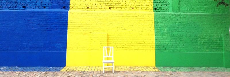 Wooden chair on footpath against multi colored brick wall