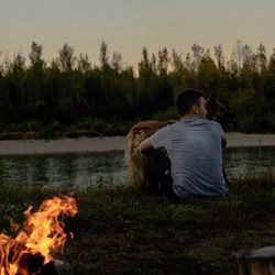 Rear view of man sitting with dog by bonfire while looking away during sunset