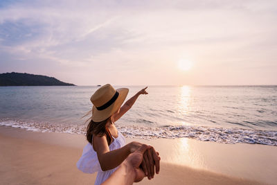 Cropped image of man holding woman hand at beach against sky during sunset