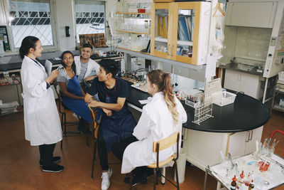 Full length of mature teacher explaining to young multi-ethnic university students sitting in chemistry laboratory