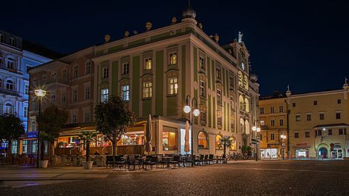 Illuminated street by buildings against sky at night