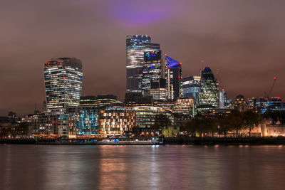 Illuminated buildings by river against sky at night