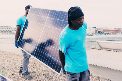 African american workers walking with solar panel on sunny day in industrial area