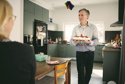 Woman looking at senior man holding birthday cake while walking by table