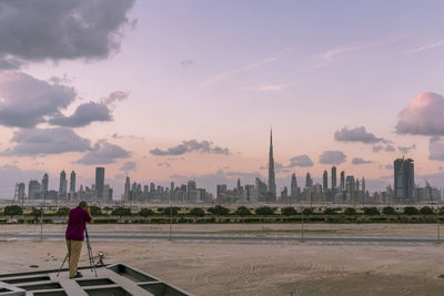Rear view of man photographing urban skyline from beach at sunset