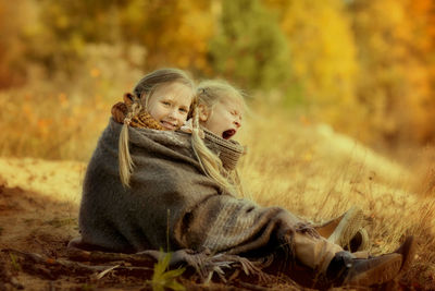 Lovely sisters are sitting, wrapped in a warm blanket, in an autumn park among the golden trees 