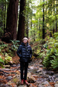 Boy with glasses and puffy coat standing on river stones in redwoods