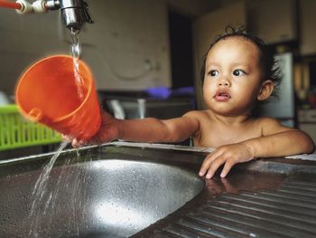 Cute baby boy washing plastic container in kitchen sink at home