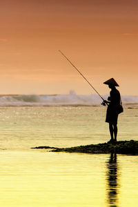 Silhouette man fishing in sea against sunset sky