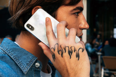 Closeup side view of crop young male with tattoo on hand having conversation on mobile phone